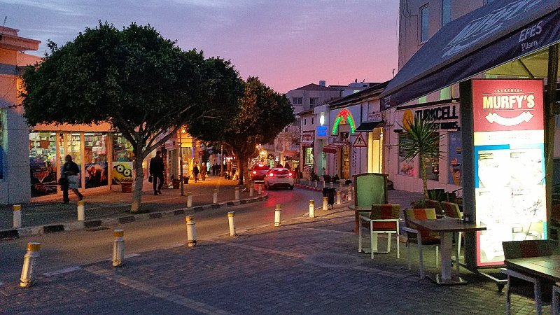 Gatebilde fra den enveiskjørte sentrumsgaten i Kyrenia med tosa solnedgangshimmel og lys i butikker. Ikke den beste restaurant Nord Kypros men populær blant unge.